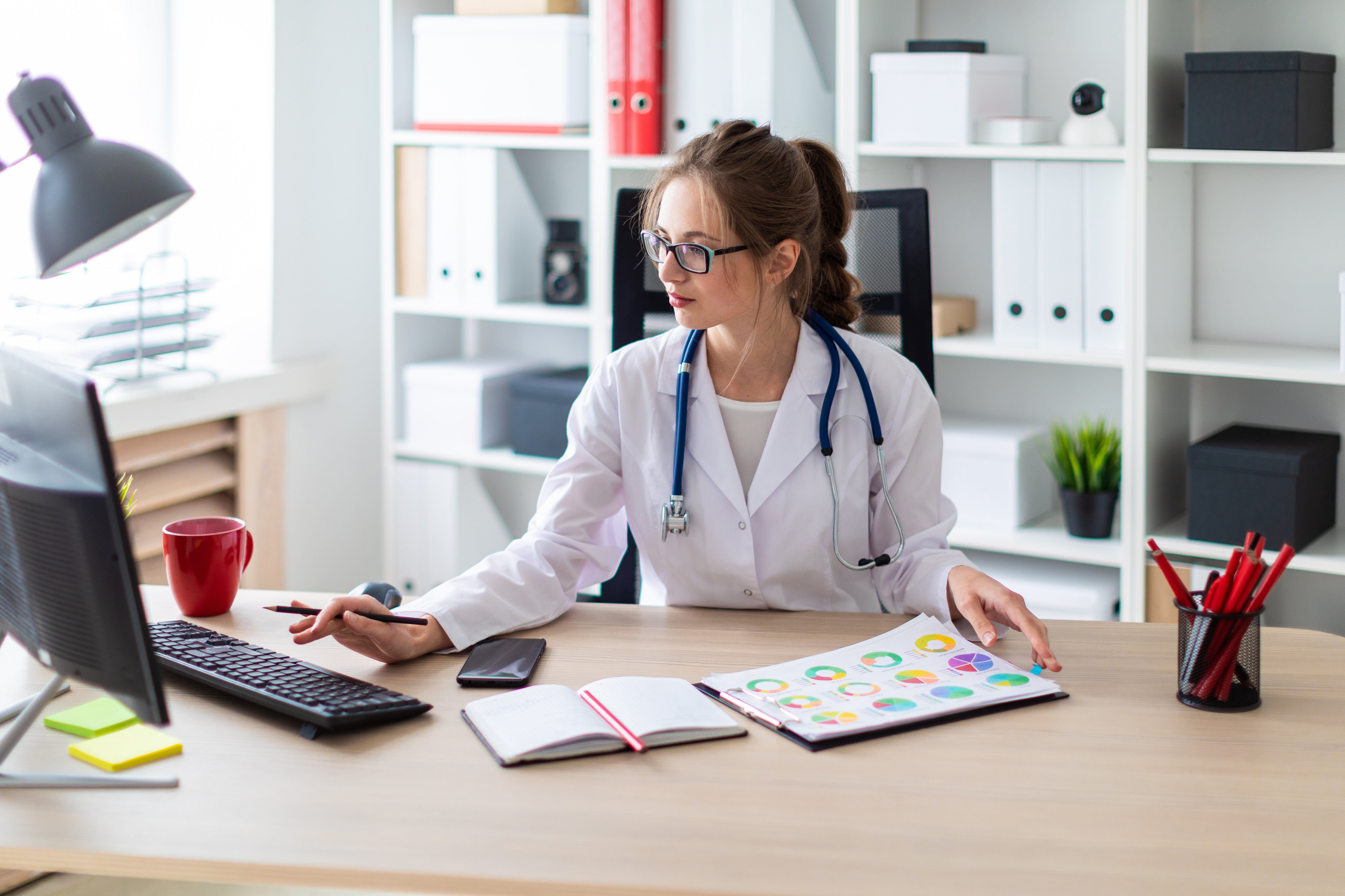 A-young-girl-in-a-white-robe-is-sitting-at-a-computer-desk,-holding-a-pencil-in-her-hand-and-working-with-a-notebook-and-charts.-1011251062_6720x4480